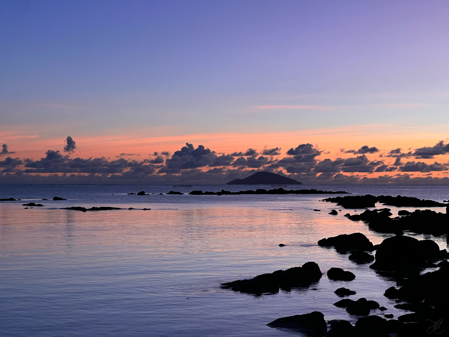 Hues of purples, lilac and faint blues with hints of remaining pinks and oranges skimming the horizon. Dark rocks create stepping stones into the calm purple waters over to the island out at sea, while foliage in the foreground adds texture and depth.