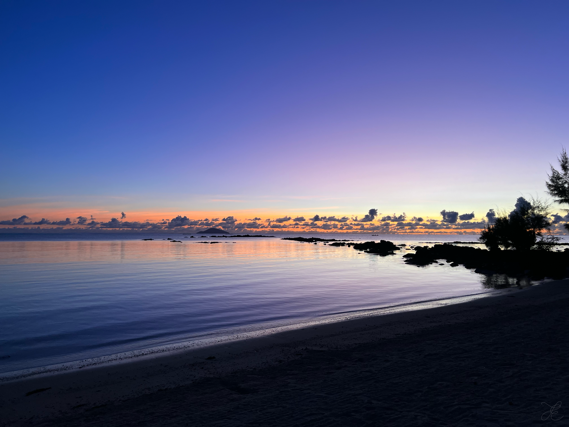 Hues of purples, lilac and faint blues with hints of remaining pinks and oranges skimming the horizon. Dark rocks create stepping stones into the calm purple waters over to the island out at sea, while foliage in the foreground adds texture and depth.