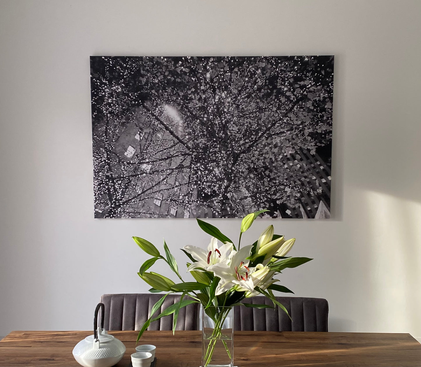Abstract monochrome photographic canvas print of tree branches illuminated by glowing fairy lights at night, blurring the New York architecture in the background.  Displayed on a white wall above dining table with a glass vase of lilies, white teapot, with sun streaming through from the windows.