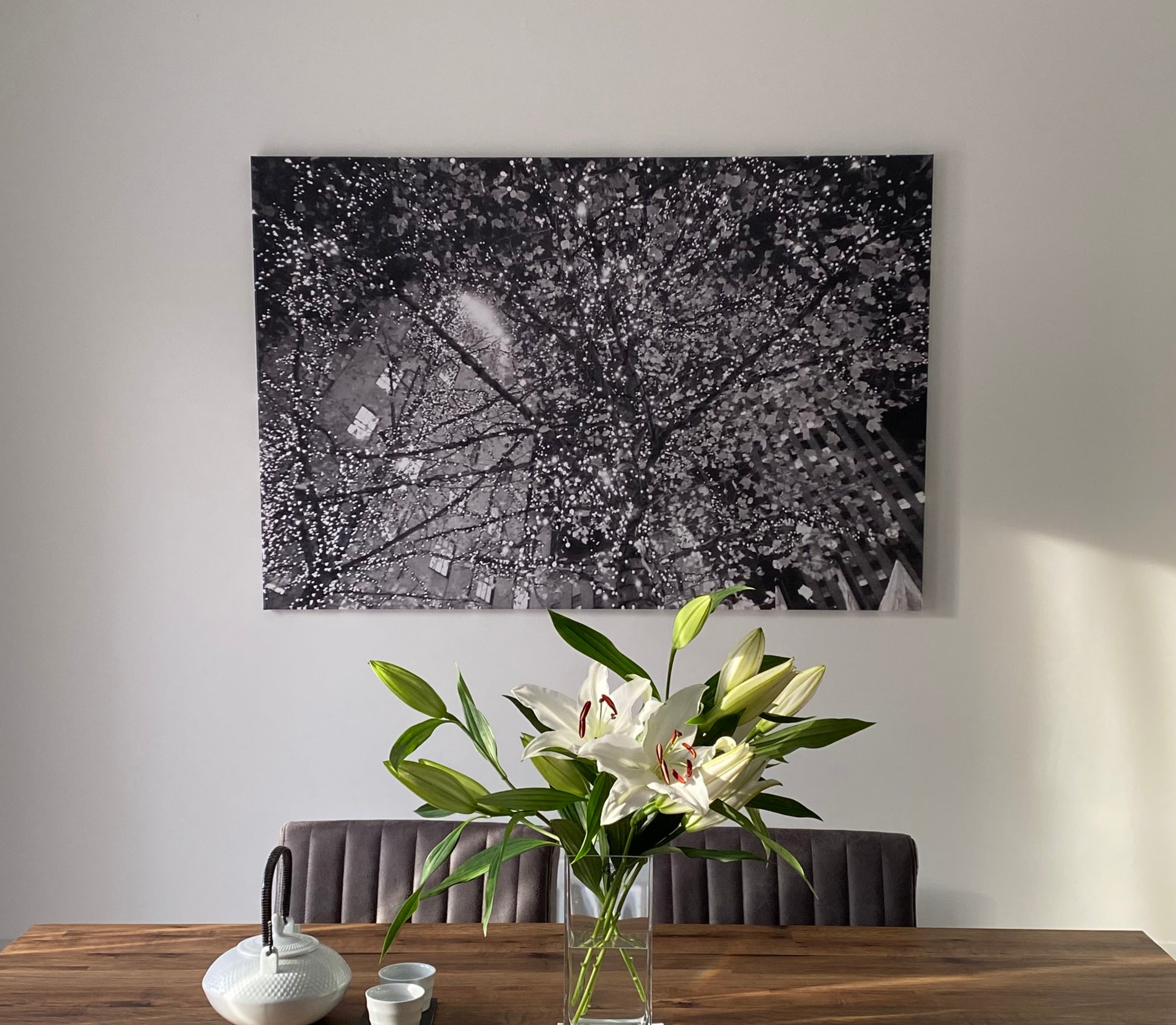 Abstract monochrome photographic canvas print of tree branches illuminated by glowing fairy lights at night, blurring the New York architecture in the background.  Displayed on a white wall above dining table with a glass vase of lilies, white teapot, with sun streaming through from the windows.
